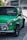 A classic, green Mini Cooper is parked on a city street, with its glossy black trim and chrome accents reflecting the ambient light. The vehicle's distinctive round headlights and compact design are prominent against the urban backdrop.