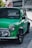 A classic, green Mini Cooper is parked on a city street, with its glossy black trim and chrome accents reflecting the ambient light. The vehicle's distinctive round headlights and compact design are prominent against the urban backdrop.