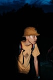 A person wearing a tan bucket hat and a yellow t-shirt with a graphic design stands against a dark background. They have a backpack with visible straps and appear to be illuminated by a direct light source, highlighting their features amidst the shadows.