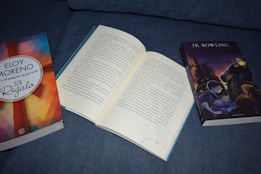 Three books are placed on a blue fabric surface. On the left, a book with a colorful geometric cover design titled 'El Regalo' by Eloy Moreno. In the center, an open book with Spanish text visible. On the right, a book featuring an illustrated cover with a wizard and castle, titled by J.K. Rowling.