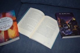 Three books are placed on a blue fabric surface. On the left, a book with a colorful geometric cover design titled 'El Regalo' by Eloy Moreno. In the center, an open book with Spanish text visible. On the right, a book featuring an illustrated cover with a wizard and castle, titled by J.K. Rowling.