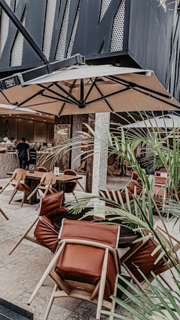 An outdoor restaurant seating area with modern decor. There are tables with chairs that have leather cushions, and a large umbrella overhead. Decorative plants are scattered around the space, and in the background, there is a counter area where people are being served.