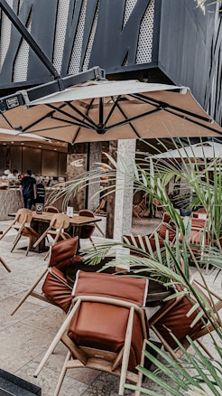 An outdoor restaurant seating area with modern decor. There are tables with chairs that have leather cushions, and a large umbrella overhead. Decorative plants are scattered around the space, and in the background, there is a counter area where people are being served.