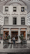 A stylish café entrance with a white facade, featuring the name 'Café' written in lights. Tall potted plants flank the doorway, and two people are standing just outside. The interior appears warm and inviting, with cozy lighting and visible seating. The glass windows reflect an intricate ceiling structure above.