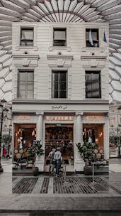 A stylish café entrance with a white facade, featuring the name 'Café' written in lights. Tall potted plants flank the doorway, and two people are standing just outside. The interior appears warm and inviting, with cozy lighting and visible seating. The glass windows reflect an intricate ceiling structure above.