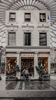 A stylish café entrance with a white facade, featuring the name 'Café' written in lights. Tall potted plants flank the doorway, and two people are standing just outside. The interior appears warm and inviting, with cozy lighting and visible seating. The glass windows reflect an intricate ceiling structure above.