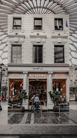 A stylish café entrance with a white facade, featuring the name 'Café' written in lights. Tall potted plants flank the doorway, and two people are standing just outside. The interior appears warm and inviting, with cozy lighting and visible seating. The glass windows reflect an intricate ceiling structure above.