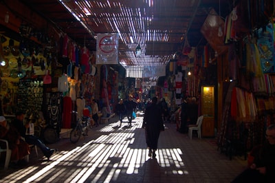 A bustling indoor marketplace with various colorful items hung on the walls, including clothing, fabrics, and decorative pieces. Overhead, a wooden lattice roof filters sunlight, casting intricate patterns on the cobbled walkway below. People are strolling through the corridor, some wearing traditional clothing, while others browse the items on display.