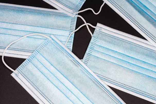 Several blue disposable surgical masks are scattered on a dark surface, showcasing their pleated, rectangular design and white elastic ear loops.