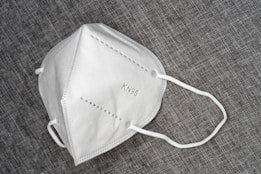A white KN95 face mask is placed on a textured gray surface. The mask features a simple design with visible stitching and ear loops.