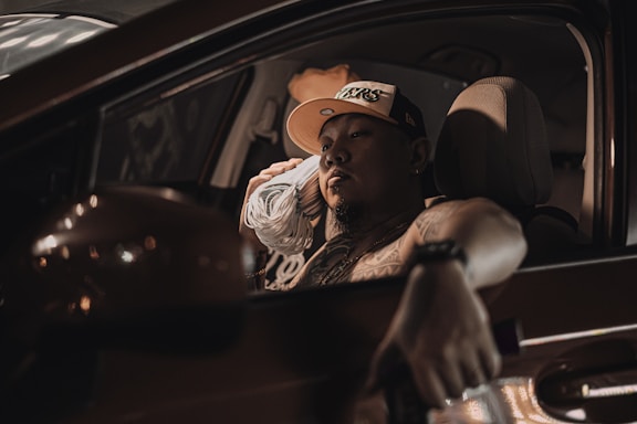 A person is sitting relaxed in the driver's seat of a car, holding what appears to be a bundle of notes or paper against their ear as if mimicking a phone conversation. The setting is dimly lit, with the car window partially open. The person is wearing a cap and appears thoughtful or contemplative.