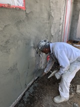 A construction worker applies cement or plaster to a rough wall surface using a trowel. The worker is dressed in white protective clothing and a cap, focused on the work. Around the construction site, there are visible pieces of construction material and the wall surface appears partially completed with areas covered in cement.