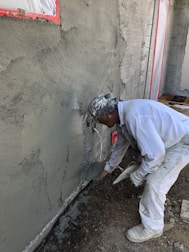 A construction worker applies cement or plaster to a rough wall surface using a trowel. The worker is dressed in white protective clothing and a cap, focused on the work. Around the construction site, there are visible pieces of construction material and the wall surface appears partially completed with areas covered in cement.