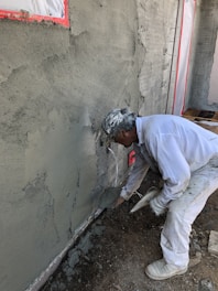 A construction worker applies cement or plaster to a rough wall surface using a trowel. The worker is dressed in white protective clothing and a cap, focused on the work. Around the construction site, there are visible pieces of construction material and the wall surface appears partially completed with areas covered in cement.