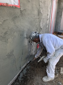 A construction worker applies cement or plaster to a rough wall surface using a trowel. The worker is dressed in white protective clothing and a cap, focused on the work. Around the construction site, there are visible pieces of construction material and the wall surface appears partially completed with areas covered in cement.