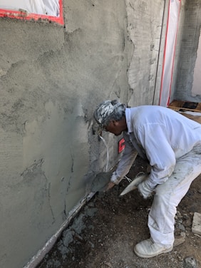 A construction worker applies cement or plaster to a rough wall surface using a trowel. The worker is dressed in white protective clothing and a cap, focused on the work. Around the construction site, there are visible pieces of construction material and the wall surface appears partially completed with areas covered in cement.