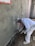 A construction worker applies cement or plaster to a rough wall surface using a trowel. The worker is dressed in white protective clothing and a cap, focused on the work. Around the construction site, there are visible pieces of construction material and the wall surface appears partially completed with areas covered in cement.