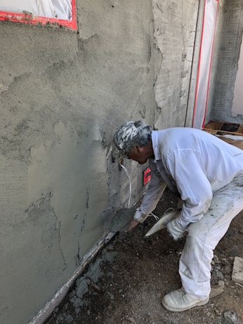 A construction worker applies cement or plaster to a rough wall surface using a trowel. The worker is dressed in white protective clothing and a cap, focused on the work. Around the construction site, there are visible pieces of construction material and the wall surface appears partially completed with areas covered in cement.