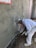 A construction worker applies cement or plaster to a rough wall surface using a trowel. The worker is dressed in white protective clothing and a cap, focused on the work. Around the construction site, there are visible pieces of construction material and the wall surface appears partially completed with areas covered in cement.