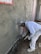 A construction worker applies cement or plaster to a rough wall surface using a trowel. The worker is dressed in white protective clothing and a cap, focused on the work. Around the construction site, there are visible pieces of construction material and the wall surface appears partially completed with areas covered in cement.