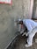 A construction worker applies cement or plaster to a rough wall surface using a trowel. The worker is dressed in white protective clothing and a cap, focused on the work. Around the construction site, there are visible pieces of construction material and the wall surface appears partially completed with areas covered in cement.