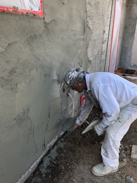 A construction worker applies cement or plaster to a rough wall surface using a trowel. The worker is dressed in white protective clothing and a cap, focused on the work. Around the construction site, there are visible pieces of construction material and the wall surface appears partially completed with areas covered in cement.