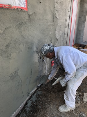 A construction worker applies cement or plaster to a rough wall surface using a trowel. The worker is dressed in white protective clothing and a cap, focused on the work. Around the construction site, there are visible pieces of construction material and the wall surface appears partially completed with areas covered in cement.