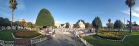 A well-maintained park with flower beds, palm trees, and benches surrounds a central fountain. Several people are walking and sitting around, enjoying the pleasant, sunny weather. Historical domed buildings and a minaret are visible in the background under a clear blue sky.