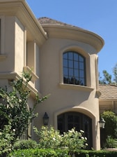 A two-story building with a smooth beige stucco exterior and rounded architectural features. Tall windows with dark frames are prominent, offering a contrast to the light facade. The roof is covered with brown tiles, and lush greenery, including bushes and trees, surrounds the structure. Decorative lantern-style wall lights are visible near the entrance.