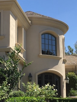 A two-story building with a smooth beige stucco exterior and rounded architectural features. Tall windows with dark frames are prominent, offering a contrast to the light facade. The roof is covered with brown tiles, and lush greenery, including bushes and trees, surrounds the structure. Decorative lantern-style wall lights are visible near the entrance.