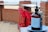 A bottle of hand sanitizer is placed on a blue plate in the foreground. It has a black pump and is positioned on a counter. Red chairs are neatly arranged in a row, backed by a brick structure. In the background, a beach with sand and water can be seen, giving the scene a casual, outdoor setting.