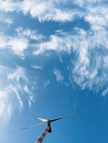 A large wind turbine is set against a bright blue sky filled with scattered, wispy clouds. The turbine's blades are positioned at an angle, and the tower features red and white stripes.