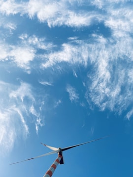 A large wind turbine is set against a bright blue sky filled with scattered, wispy clouds. The turbine's blades are positioned at an angle, and the tower features red and white stripes.