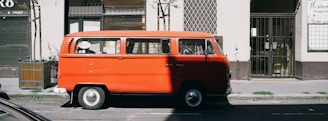 A vintage red van, likely a classic Volkswagen model, is parked on the side of the street in an urban setting. The van is in a shaded area, though sunlight highlights parts of its vibrant red exterior. Surrounding the van are elements typical of city streets, such as a storefront with a rolling shutter and a tree in a planter box. The background features a gated entrance and a patterned wall, adding to the urban atmosphere.