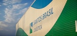 A large building features the branding 'Santos Brasil Logistica' with colorful patterns. The structure is positioned under a partly cloudy sky.