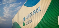 A large building features the branding 'Santos Brasil Logistica' with colorful patterns. The structure is positioned under a partly cloudy sky.
