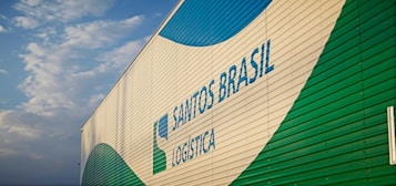 A large building features the branding 'Santos Brasil Logistica' with colorful patterns. The structure is positioned under a partly cloudy sky.