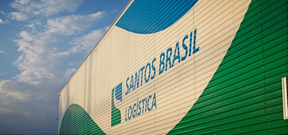 A large building features the branding 'Santos Brasil Logistica' with colorful patterns. The structure is positioned under a partly cloudy sky.