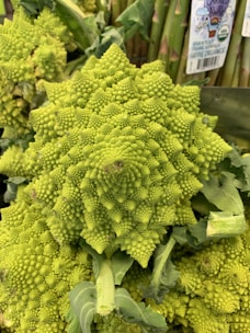 A detailed close-up of a Romanesco broccoli displaying its unique fractal patterns. The vegetable is bright green and surrounded by dark, leafy greens in the background, with some asparagus visible. There are organic labels visible on the asparagus.