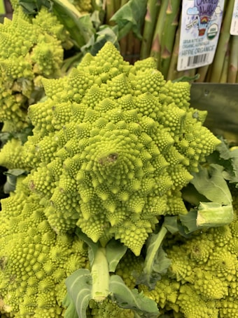 A detailed close-up of a Romanesco broccoli displaying its unique fractal patterns. The vegetable is bright green and surrounded by dark, leafy greens in the background, with some asparagus visible. There are organic labels visible on the asparagus.