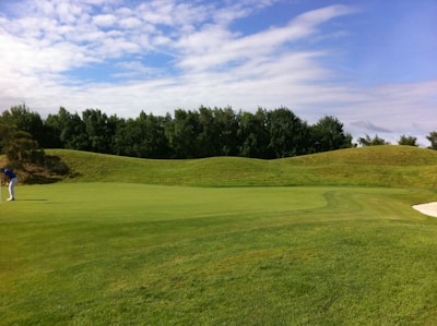 A golfer wearing a blue top and white pants is preparing to putt on a well-maintained green, surrounded by gentle grassy hills and dense trees under a partly cloudy sky.