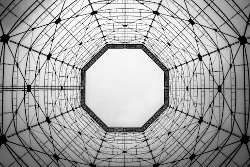 A geometric view is looking up into a steel structure with an octagonal shape. The structure consists of intersecting lines and beams, creating a symmetrical pattern. The background is a light gray, contrasting with the dark lines, highlighting the intricate architectural design.