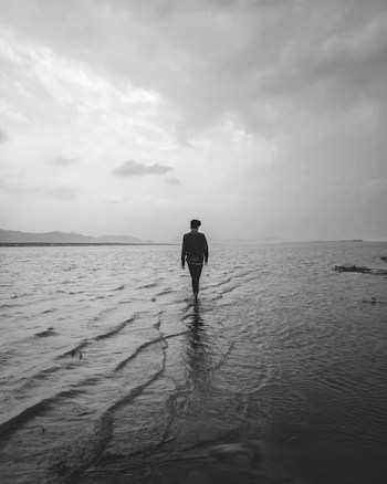 A lone figure walks along the shoreline, their reflection visible in the wet sand. The sky is overcast with scattered clouds, creating a moody and serene atmosphere. The horizon is barely visible, blending into the distant hills or mountains. The monochrome effect adds to the contemplative nature of the scene.