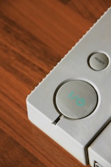 A close-up view of a gray electronic device featuring two buttons. One large circular button displays a power symbol with a light blue accent, and a smaller circular button labeled 'RESET' is situated to its side. The background has a rich, warm-toned wooden surface.