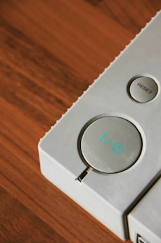 A close-up view of a gray electronic device featuring two buttons. One large circular button displays a power symbol with a light blue accent, and a smaller circular button labeled 'RESET' is situated to its side. The background has a rich, warm-toned wooden surface.