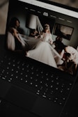 A black laptop displays an image of a bride in a wedding dress surrounded by people adjusting her veil inside a softly lit room. The room has a modern, elegant decor with a lamp and framed artwork on the wall.