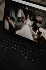 A black laptop displays an image of a bride in a wedding dress surrounded by people adjusting her veil inside a softly lit room. The room has a modern, elegant decor with a lamp and framed artwork on the wall.