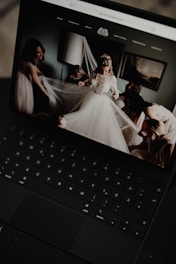 A black laptop displays an image of a bride in a wedding dress surrounded by people adjusting her veil inside a softly lit room. The room has a modern, elegant decor with a lamp and framed artwork on the wall.