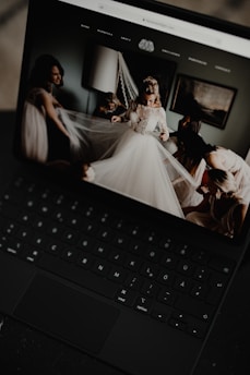 A black laptop displays an image of a bride in a wedding dress surrounded by people adjusting her veil inside a softly lit room. The room has a modern, elegant decor with a lamp and framed artwork on the wall.