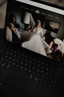 A black laptop displays an image of a bride in a wedding dress surrounded by people adjusting her veil inside a softly lit room. The room has a modern, elegant decor with a lamp and framed artwork on the wall.